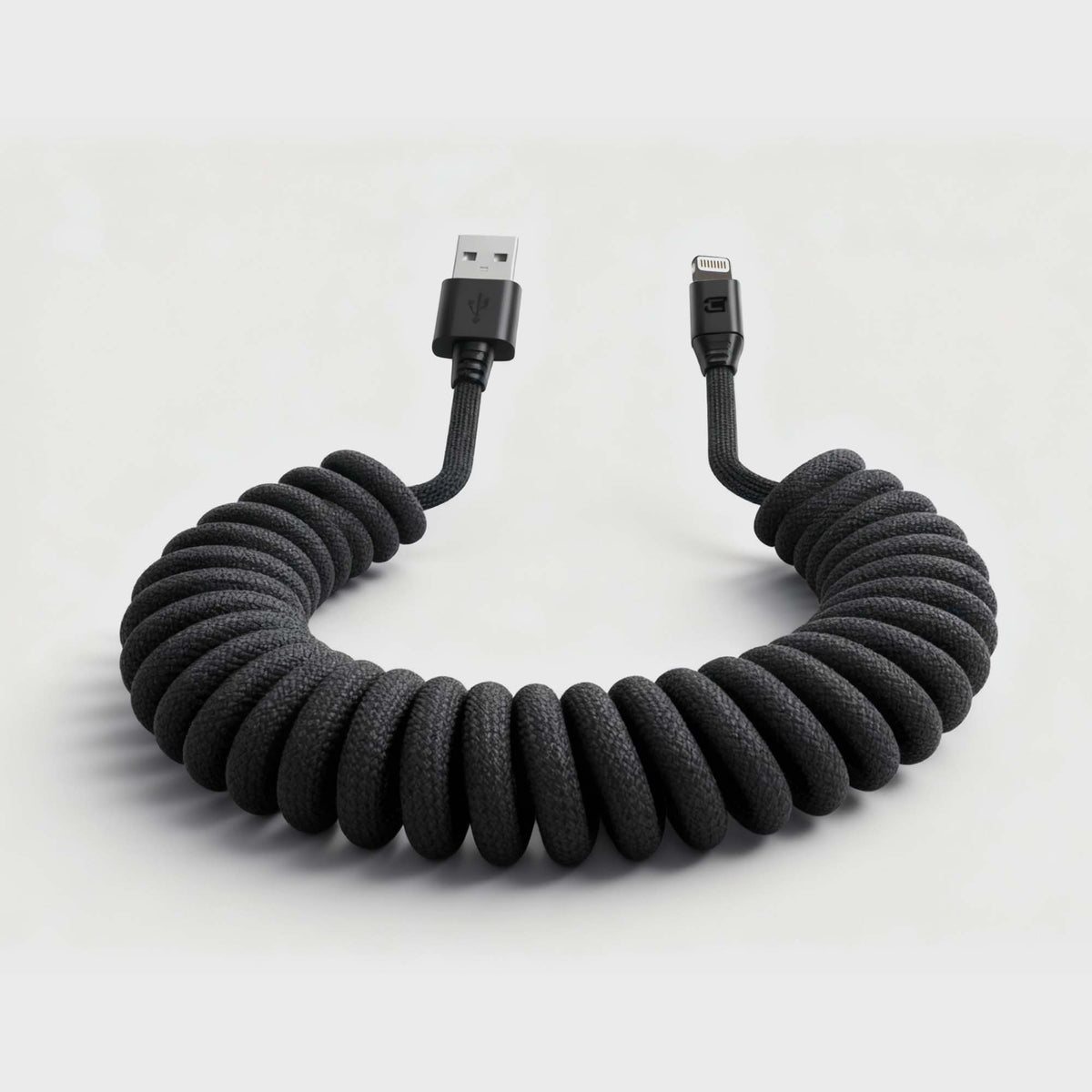 Black braided coiled USB-A to Lightning cable with magnetic connectors, lying on a light-colored desk, emphasizing durability and style